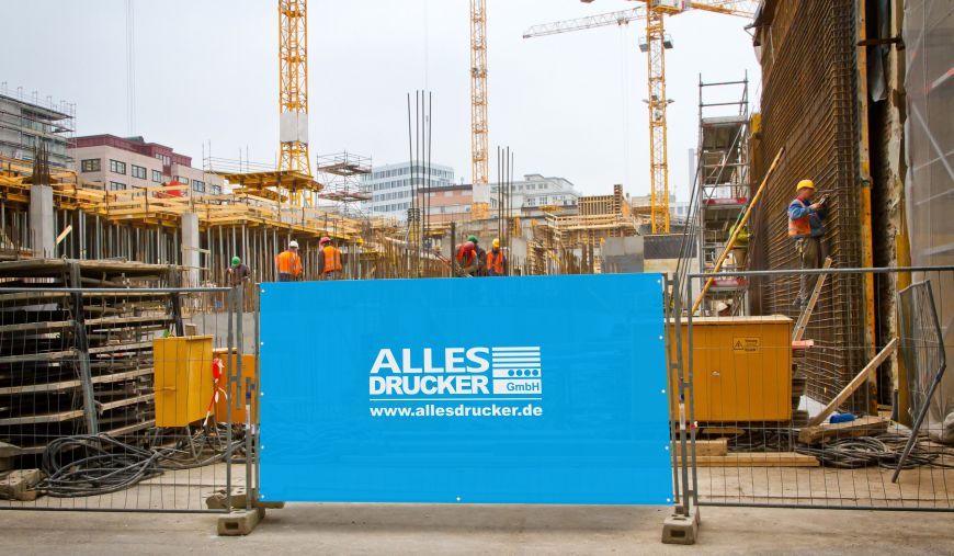 Blaue Bauzaun Husse mit dem Allesdrucker Logo vor einer Baustelle